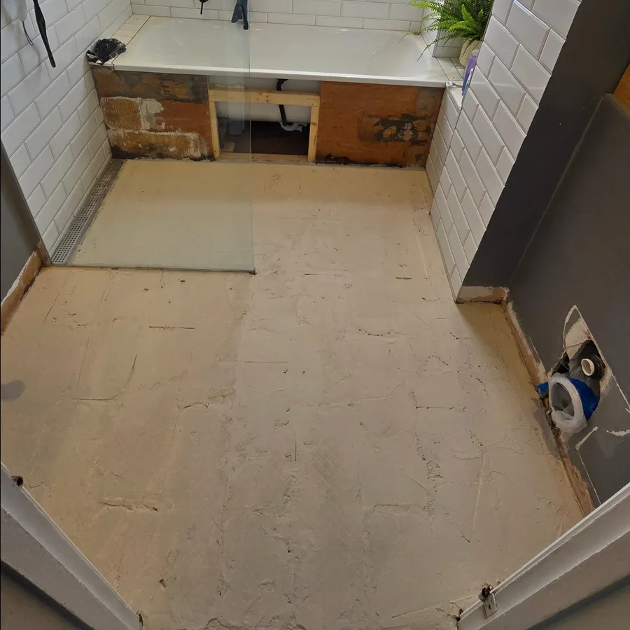 Bathroom floor preparation surface with a linear drain beside white tiled walls.